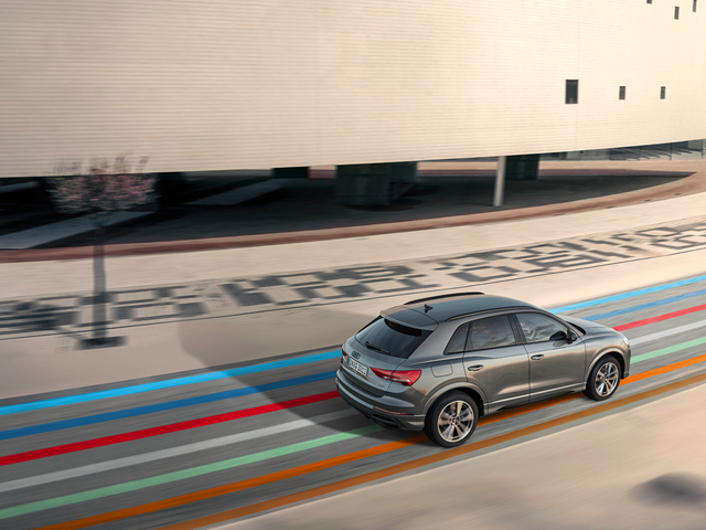 Audi Q3 fährt durch eine moderne Stadtstraße mit buntem Hahn Code.