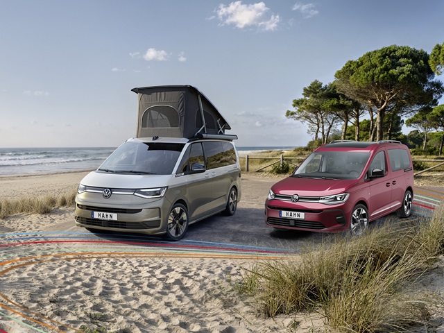 Roter VW Caddy und grauer California mit offenem Aufstelldach auf einem Küstenparkplatz mit Sanddünen und Meer im Hintergrund.