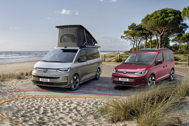 Roter VW Caddy und grauer California mit offenem Aufstelldach auf einem Küstenparkplatz mit Sanddünen und Meer im Hintergrund.