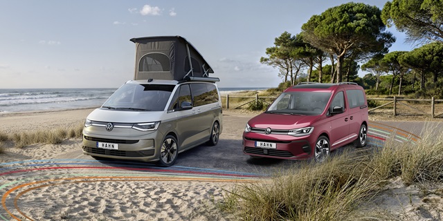Roter VW Caddy und grauer California mit offenem Aufstelldach auf einem Küstenparkplatz mit Sanddünen und Meer im Hintergrund.