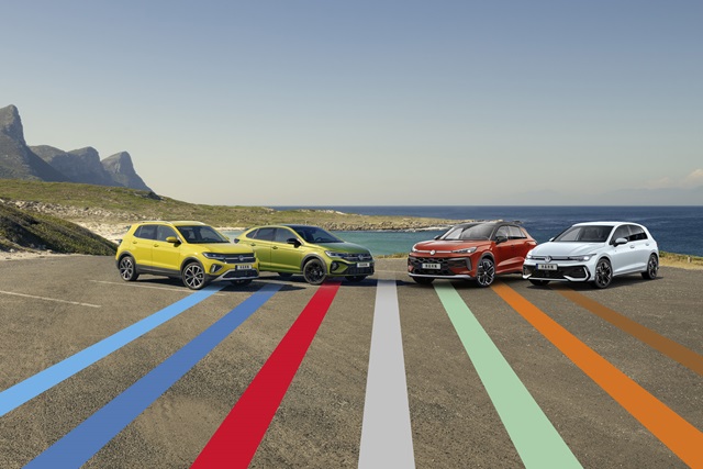 Vier verschiedene Volkswagen-Modelle nebeneinander auf einer Küstenstraße, im Hintergrund Meer und Berge.