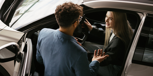 Frau sitzt am Steuer eines Autos und spricht mit einem Mann, der außen am geöffneten Fenster steht