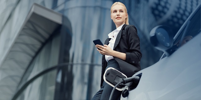 Frau in Businesskleidung steht neben einem Elektroauto und hält ein Smartphone in der Hand.
