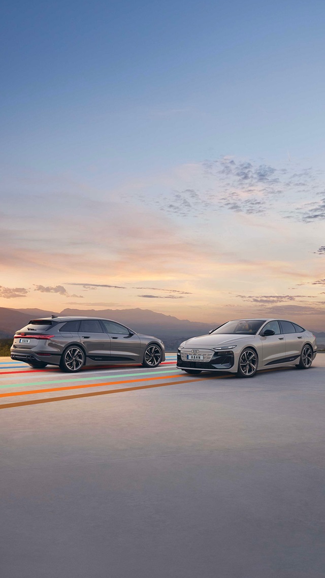 Zwei Audi A6 e-tron Fahrzeuge, ein Kombi und eine Limousine, stehen nebeneinander auf einer glatten Fläche vor einem Himmel mit leicht bewölktem Sonnenuntergang.