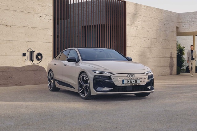 Beige Audi A6 e-tron Sportback in Frontansicht vor moderner Fassade mit Ladestation an der Wand und Person im Hintergrund