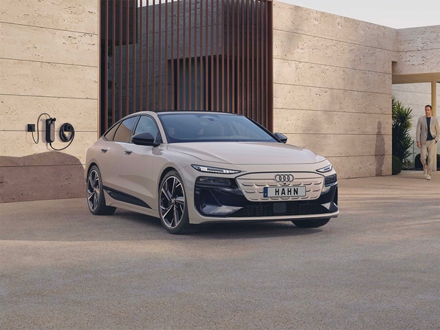 Beige Audi A6 e-tron Sportback in Frontansicht vor moderner Fassade mit Ladestation an der Wand und Person im Hintergrund
