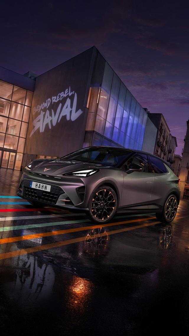 Der CUPRA Raval steht vor einem modernen Glasgebäude mit der Projektion 'PROUD REBEL RAVAL' auf der Fassade, das Auto hat ein grau-lila Finish und schwarze Felgen mit orangefarbenen Akzenten.