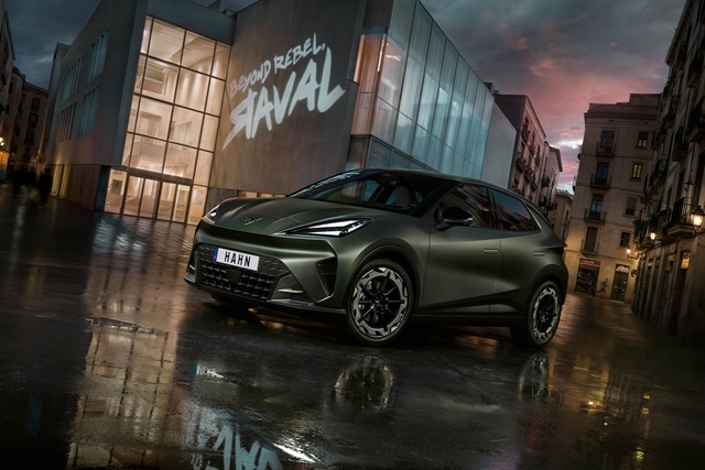 Cupra Raval VZ Extreme in mattgrüner Lackierung steht vor einem modernen Gebäude mit beleuchteter Glasfassade und Schriftzug 'BEYOND REBEL RAVAL' bei Dämmerung auf nassem Boden.
