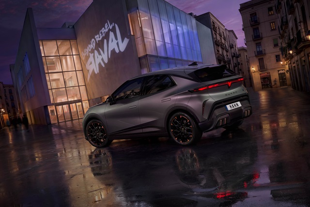 Heckansicht des grauen CUPRA Raval VZ Extreme vor einem modernen Gebäude mit beleuchtetem Schriftzug 'SOUND REBEL RAVAL' bei Dämmerung auf nassem Pflaster.