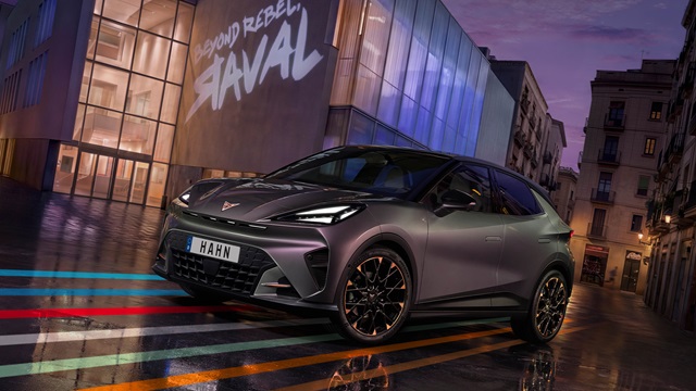 Der CUPRA Raval steht vor einem modernen Glasgebäude mit der Projektion 'PROUD REBEL RAVAL' auf der Fassade, das Auto hat ein grau-lila Finish und schwarze Felgen mit orangefarbenen Akzenten.