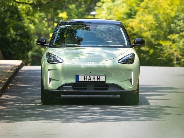 Geely E2 in mintgrün in der Frontansicht fährt auf einer Straße und im Hintergrund sind grüne Bäume