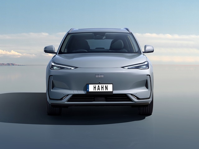 Grauer Geely E5 in Frontansicht mit schmalen Scheinwerfern und sichtbarem Kennzeichen mit dem Schriftzug 'HAHN' auf leerer Fläche.