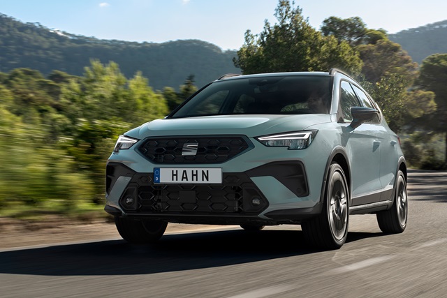 Frontansicht des grauen SEAT Arona SUV mit schwarzem Dach und markantem Kühlergrill auf einer Straße vor bewaldetem Hintergrund