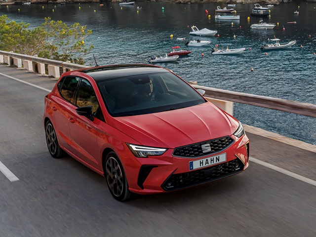 Roter SEAT Ibiza fährt auf Küstenstraße mit Wasser und Booten im Hintergrund
