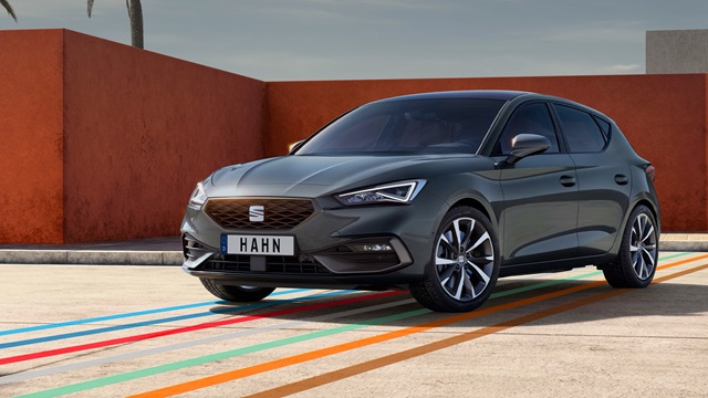 SEAT Leon in grauer Lackierung steht vor einer orangefarbenen Wand mit Palmen im Hintergrund auf einem Parkplatz mit farbigen Linien auf dem Boden