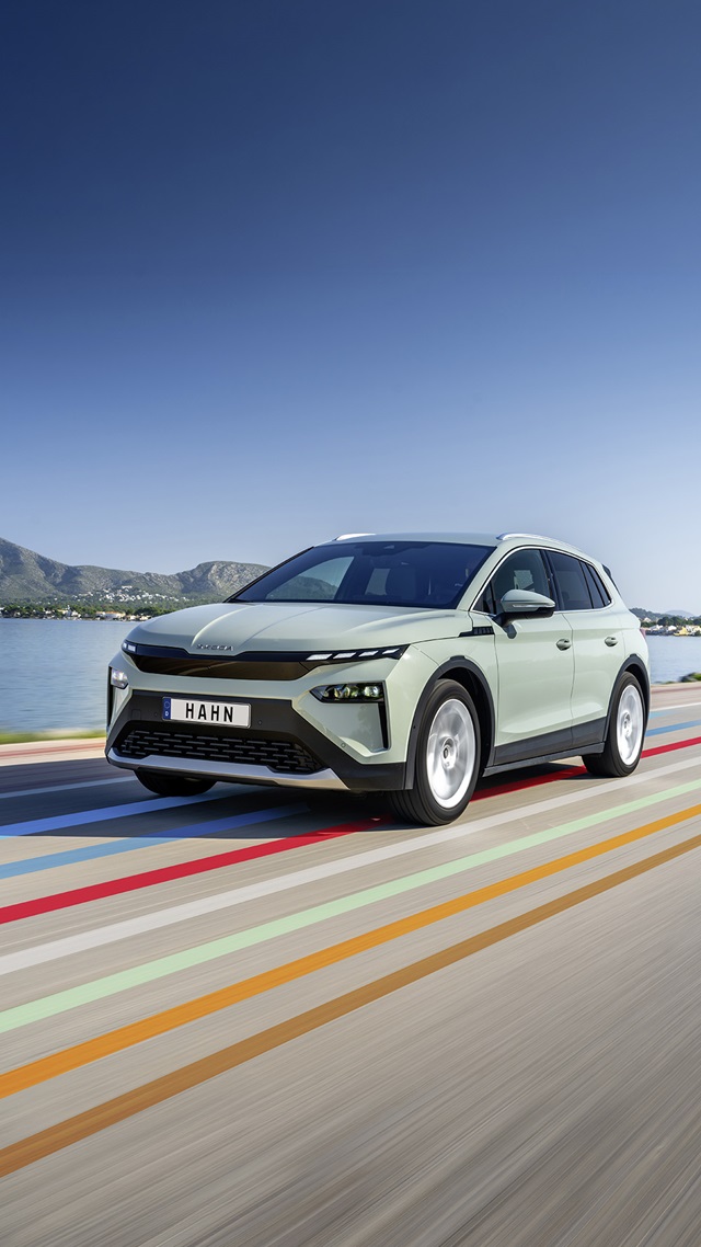 Skoda Elroq SUV in grüner Lackierung fährt auf einer Straße mit bunten Streifen vor einem See und Bergen im Hintergrund