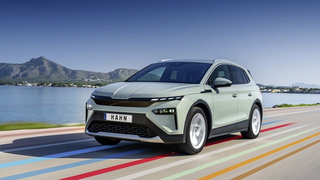 Skoda Elroq SUV in grüner Lackierung fährt auf einer Straße mit bunten Streifen vor einem See und Bergen im Hintergrund