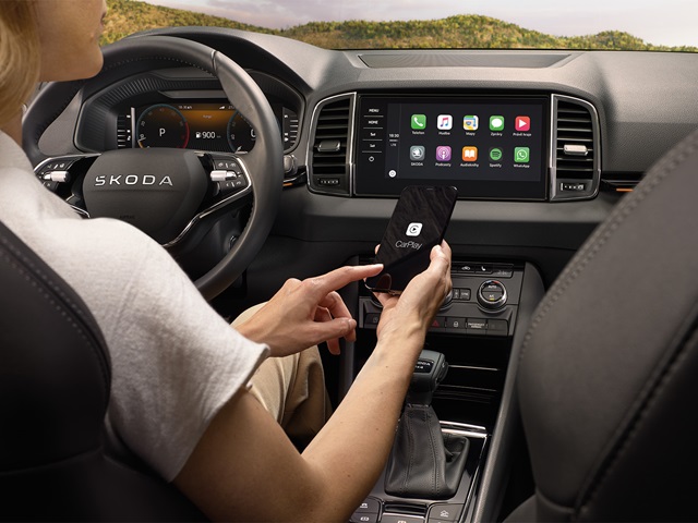 Fahrerin im Škoda Karoq tippt auf ein Smartphone mit CarPlay-Logo vor dem zentralen Touchscreen mit App-Symbolen.
