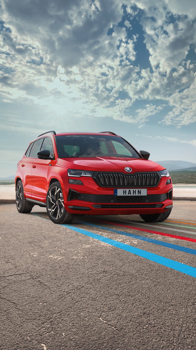 Ein roter Skoda Karoq SUV steht auf einer asphaltierten Straße vor einem bewölkten Himmel, mit farbigen Linien auf dem Boden und HAHN-Kennzeichen.