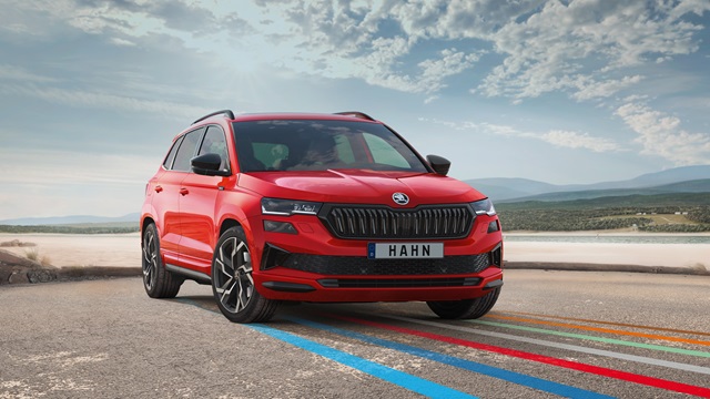 Ein roter Skoda Karoq SUV steht auf einer asphaltierten Straße vor einem bewölkten Himmel, mit farbigen Linien auf dem Boden und HAHN-Kennzeichen.