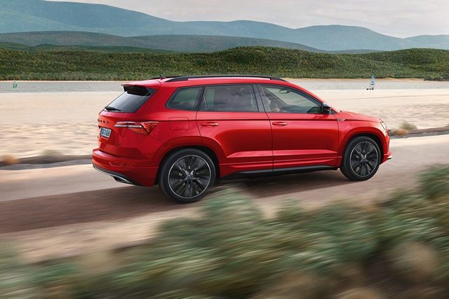 Ein roter Skoda Karoq SUV fährt auf einer Küstenstraße entlang, mit Bergen im Hintergrund und einer unscharfen Vordergrundlandschaft durch die Fahrt.
