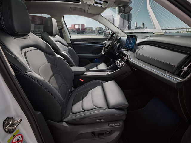 Innenraum des Škoda Kodiaq mit zwei schwarzen Vordersitzen, einem mittig angeordneten Armaturenbrett mit großem Touchscreen und einem schwarzen Lenkrad vor einer urbanen Flusslandschaft mit Brücke