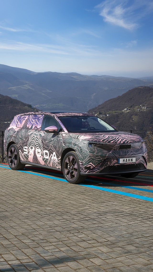 Škoda Peaq Erlkönig mit auffälliger Camouflage-Folie steht auf gepflastertem Parkplatz vor Berglandschaft.
