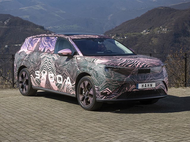 Škoda Peaq Erlkönig mit auffälliger Camouflage-Folie steht auf gepflastertem Parkplatz vor Berglandschaft.