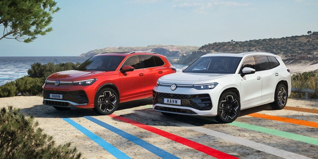 Zwei SUV nebeneinander in Seitenansicht auf einer Straße mit farbigen Streifen, links ein roter VW Tayron, rechts ein weißer VW Tiguan, im Hintergrund Meer und Hügel