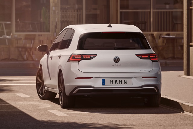 Weiße Heckansicht eines VW Golf E-Hybrid mit deutschem Kennzeichen 'HAHN' auf einer Straße vor unscharfem urbanem Hintergrund