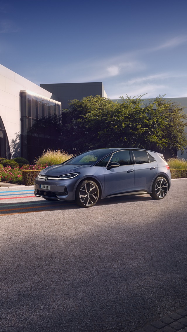 Seitliche Ansicht eines blauen VW ID.3 Neo vor einem modernen Gebäude mit Büschen und Blumen im Hintergrund