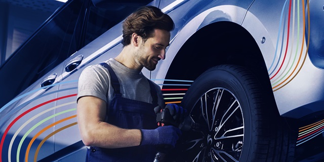 Mann in blauer Arbeitskleidung montiert mit einem Werkzeug einen Reifen an einem Auto.