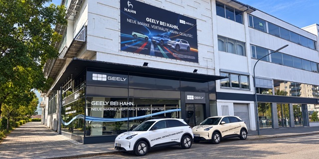 Zwei Geely E2 Modelle stehen vor einem modernen, mehrstöckigen Gebäude mit großen Fenstern und einem Werbebanner für Geely bei Hahn in Pforzheim